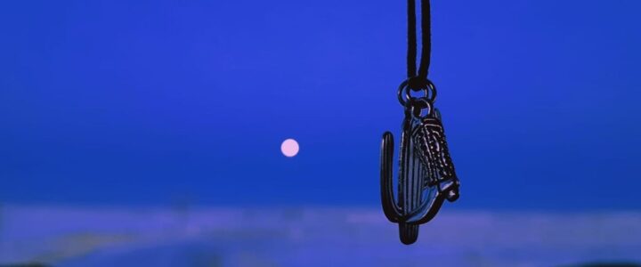 Médaillons en forme de cactus de de botte de cowboy suspendu devant un parebrise de camion avec en arrière-plan la lune rose sur un ciel bleu foncé.