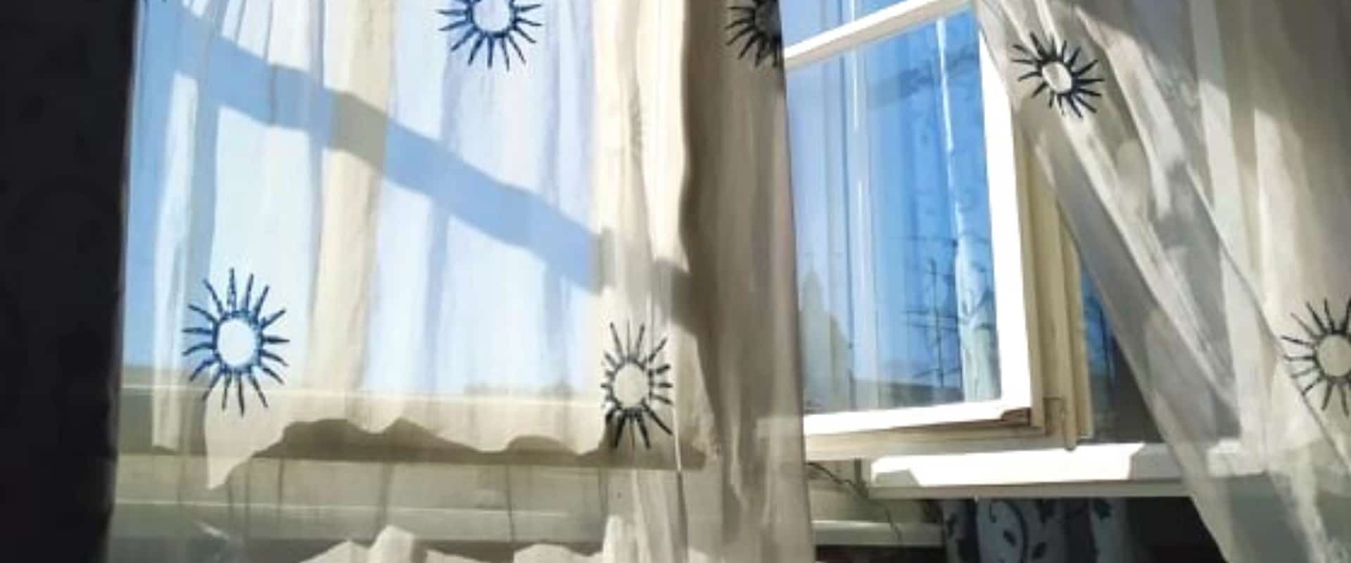 Rideau blanc diaphane avec soleils brodés en bleu ballotant au vent devant une fenêtre ouverte.