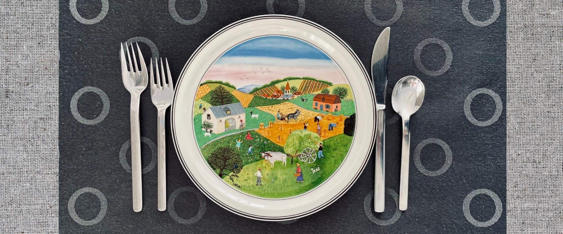 Un couvert sur une table incluant une assiette décorée par une scène de champs cultivés.