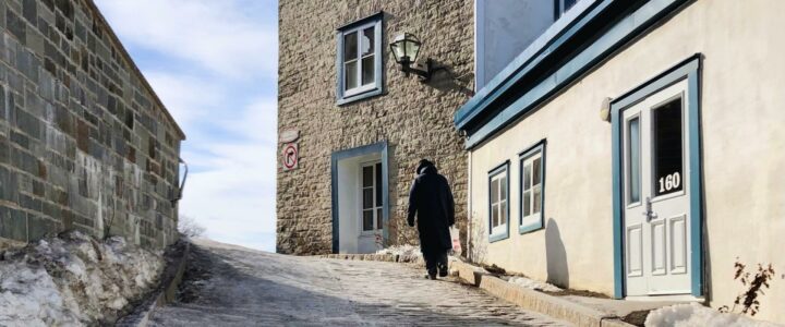 Un homme d’un certain âge portant un pardessus foncé vu de dos marche dans la rue.