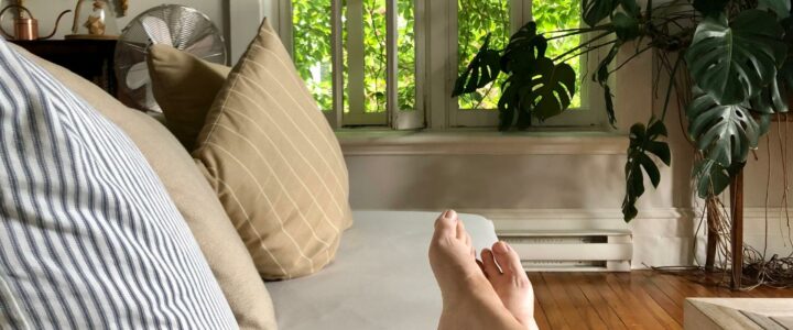 Pieds étendus sur un sofa blanc devant fenêtre couvertes de plantes.